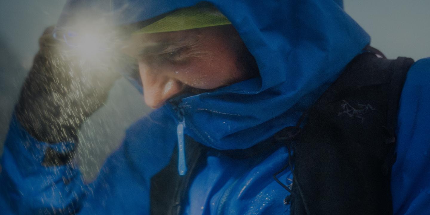 Person in a blue Arc'teryx jacket and hood, illuminated by a headlamp in falling snow or rain.