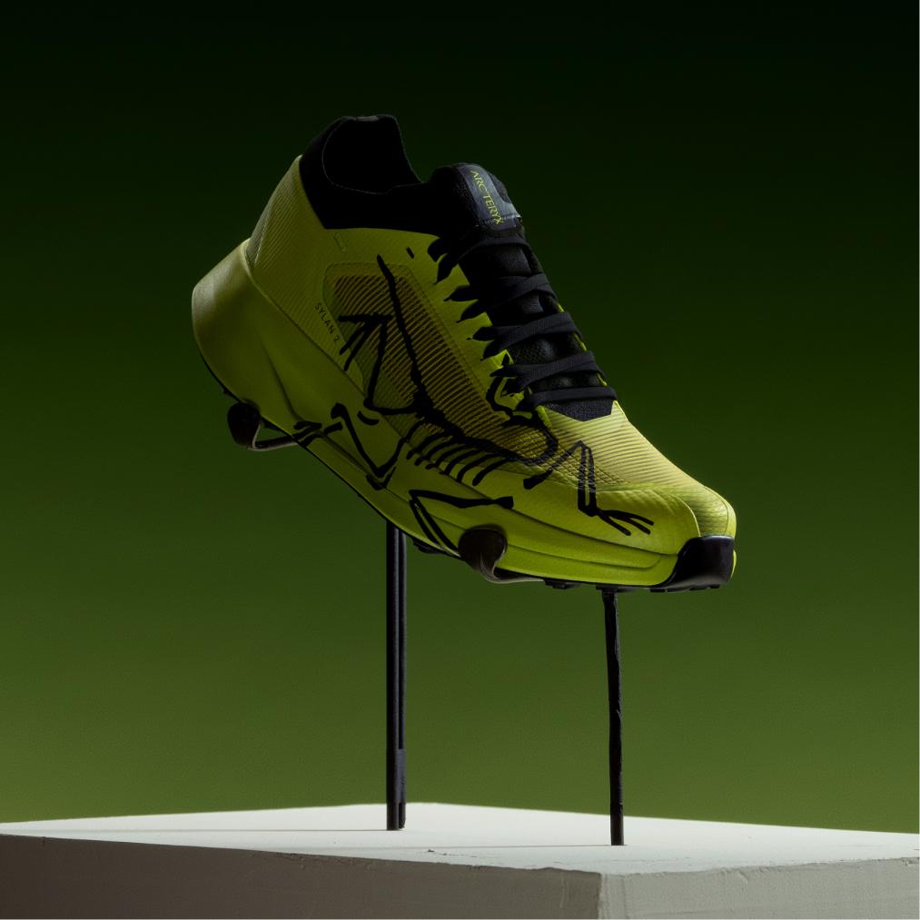 Bright yellow-green Arc'teryx Sylan 2 running shoe with a black frog skeleton design, displayed on rods against a dark green background.