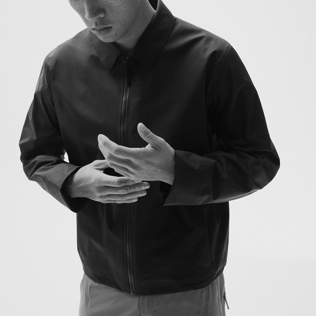 Man in a dark collared zip-up jacket, looking down with hands clasped.