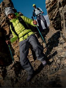 Two hikers, a woman in a bright green jacket and a man behind her, carefully descend a steep, rocky mountain path with trekking poles.