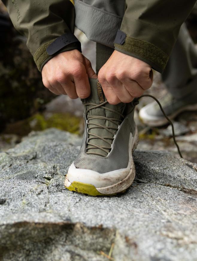 A person's hands adjusting an olive green Arc'teryx hiking boot on a rocky surface.