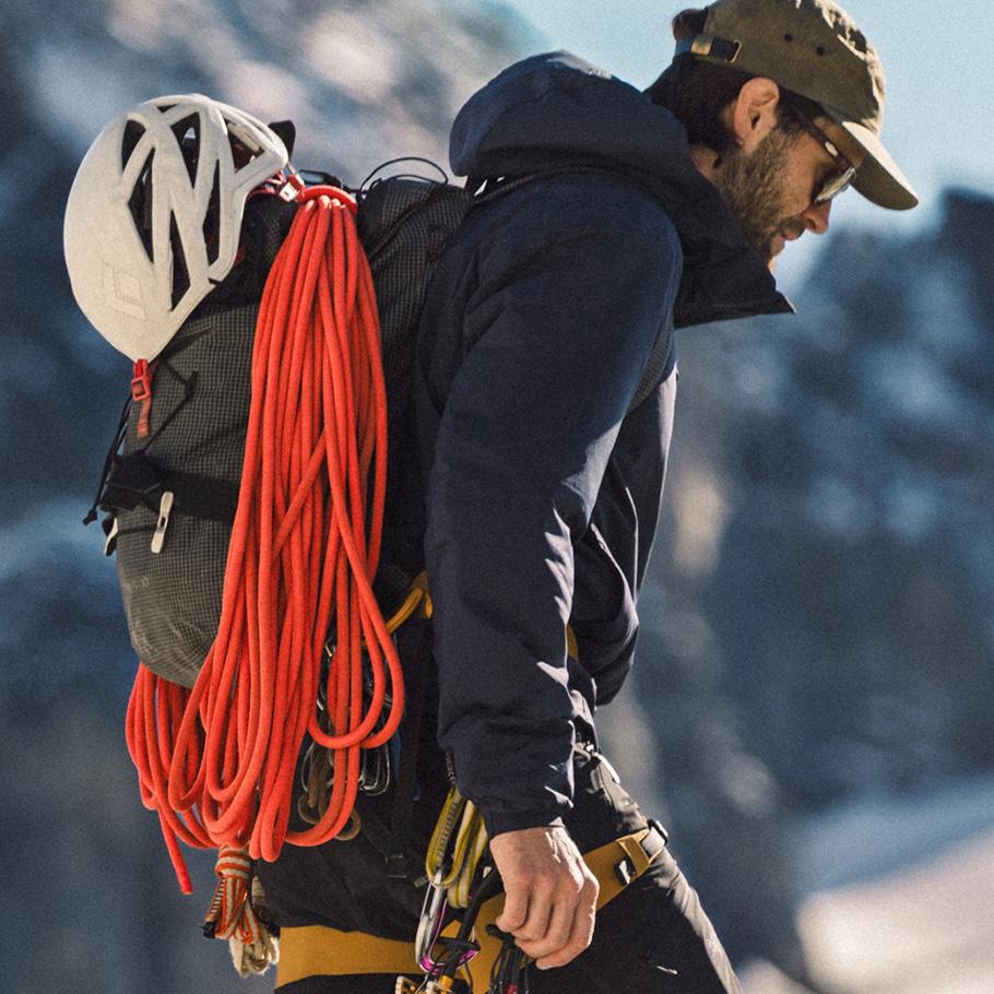 A climber in a dark jacket, cap, and sunglasses, with a white helmet, orange rope, and harness gear, looking down.