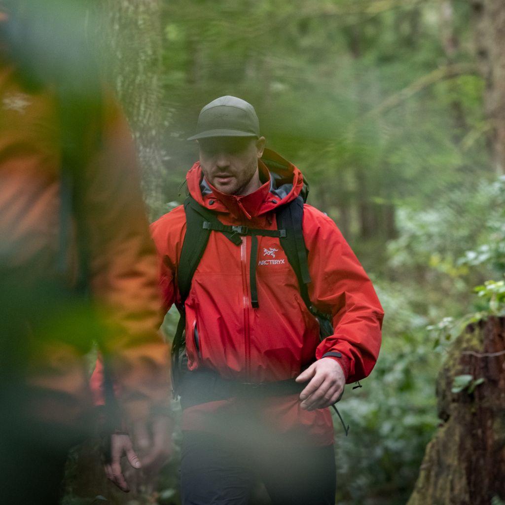 Man in a red Arc'teryx jacket and cap hiking in a forest.