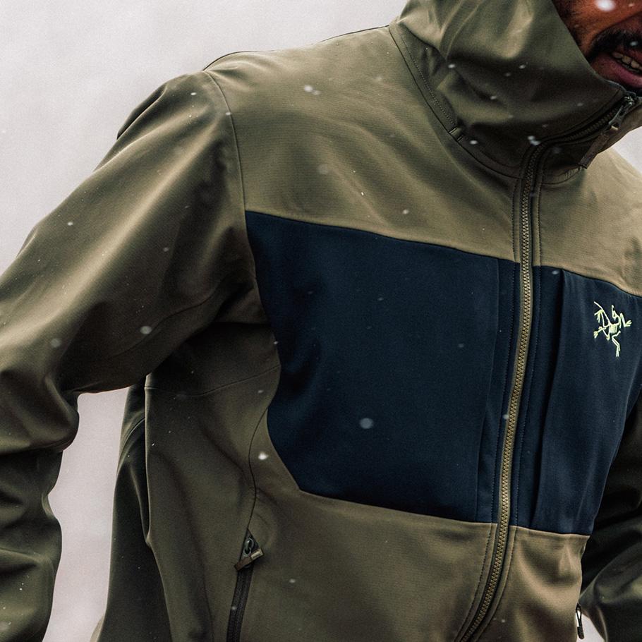 A person wearing an olive green and dark blue Arc'teryx jacket with falling snow.