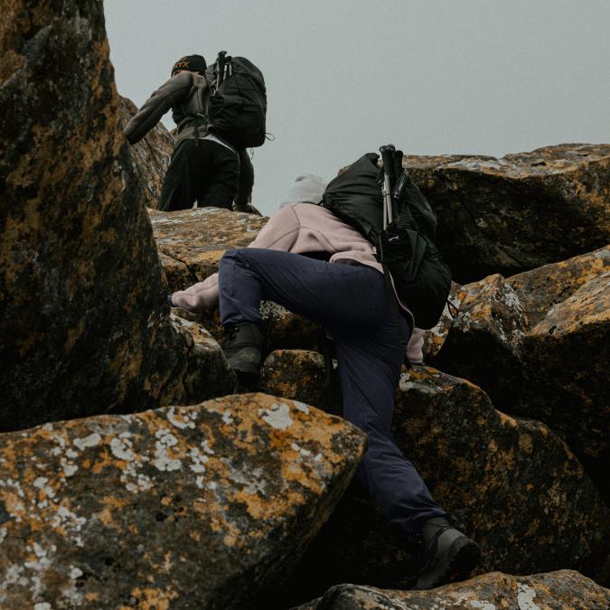 Two hikers with backpacks scramble over large, lichen-covered rocks.