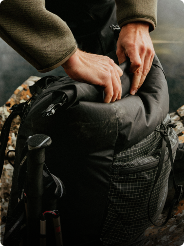 Hands closing a black roll-top hiking backpack with a trekking pole attached.