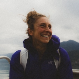 A broadly smiling woman in a purple jacket sits in a boat with mountains and water in the background.
