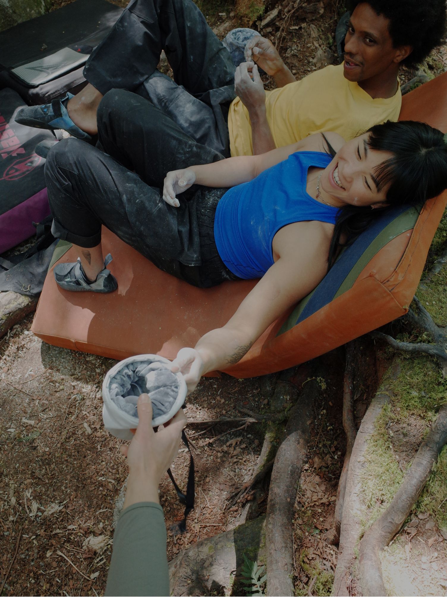Two climbers relax on crash pads while one hands a chalk bag to a smiling woman.