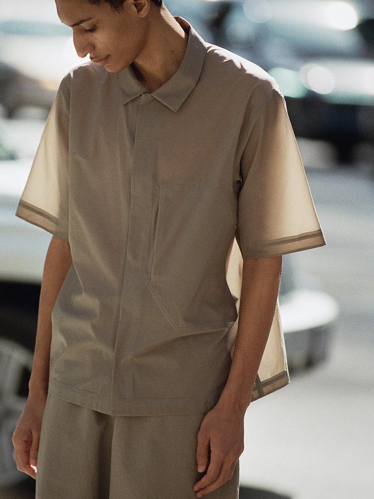 A person wears a light brown short-sleeved collared shirt with sheer, striped cuffs and matching shorts, looking down.