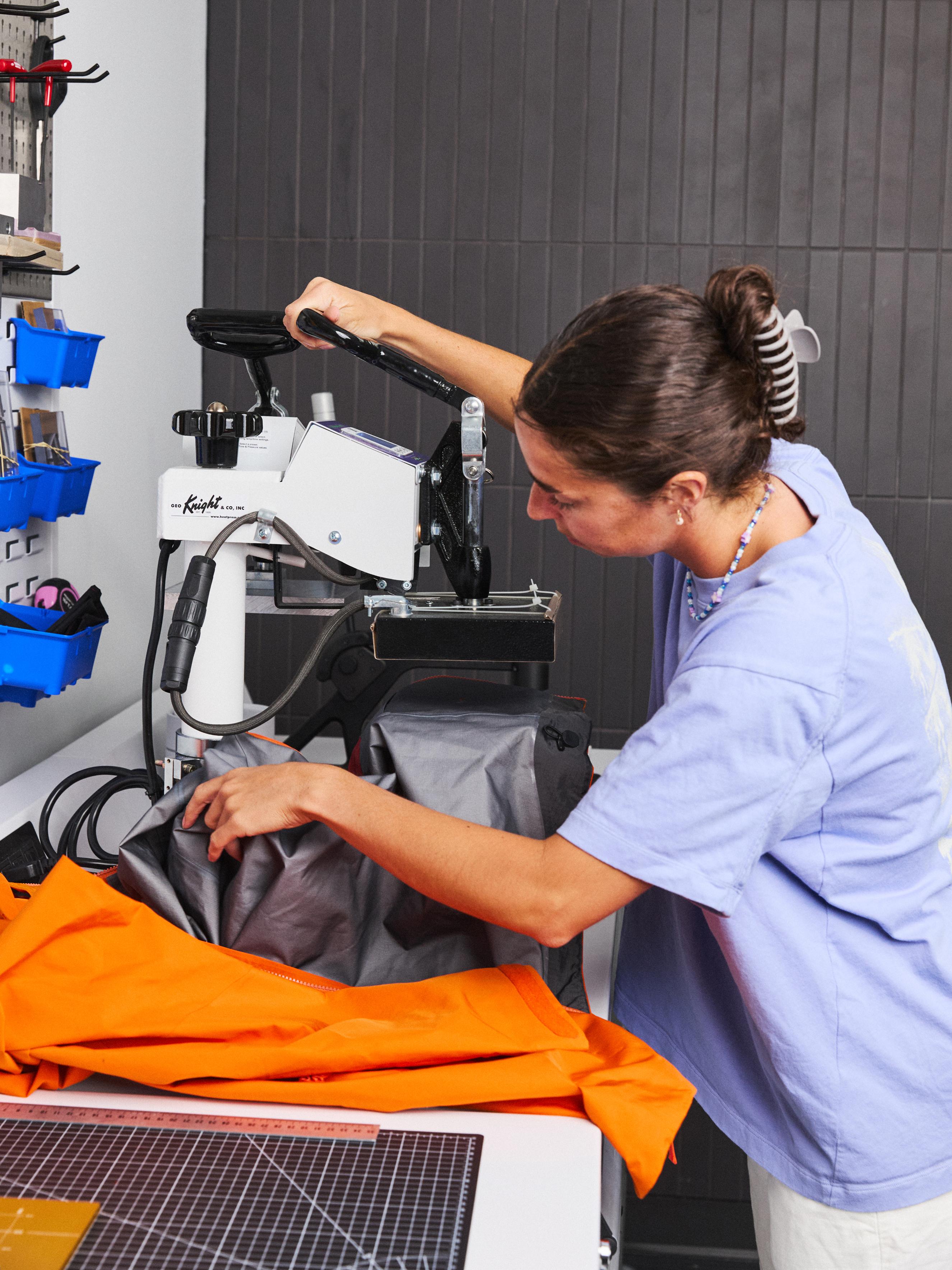 A woman operates a heat press machine, with orange fabric on the table in front of her.
