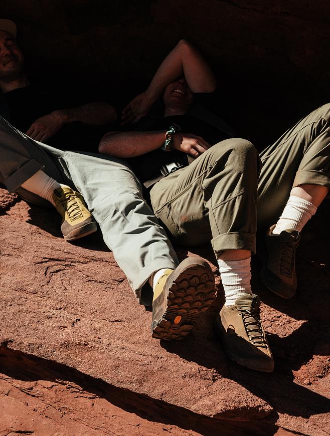 Two people wearing hiking shoes and pants relax on a red rock surface, their faces largely in shadow.
