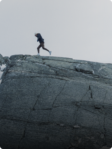 A person runs across a rocky, snow-dusted ridge under a pale sky.