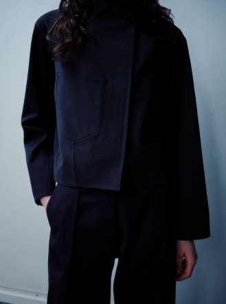 A person wears a dark blue cropped jacket with an asymmetrical front design and pocket, paired with dark pants.