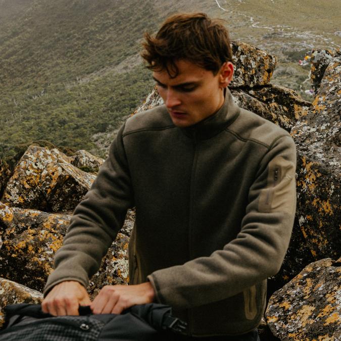 Young man in an olive fleece jacket reaching into a backpack on a rocky mountain.