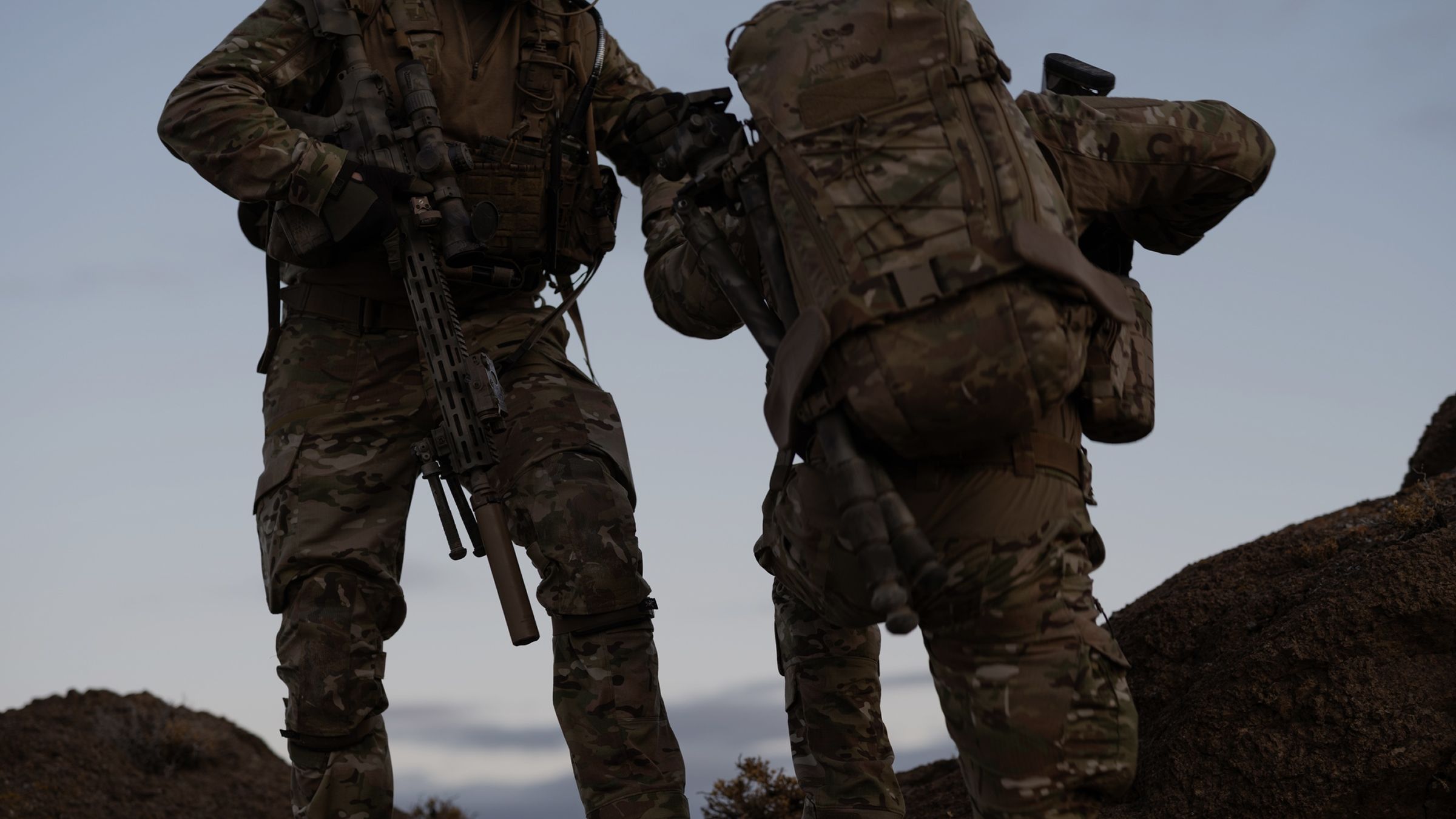 Two soldiers in camouflage uniforms and tactical gear, one assisting the other up a rocky incline.