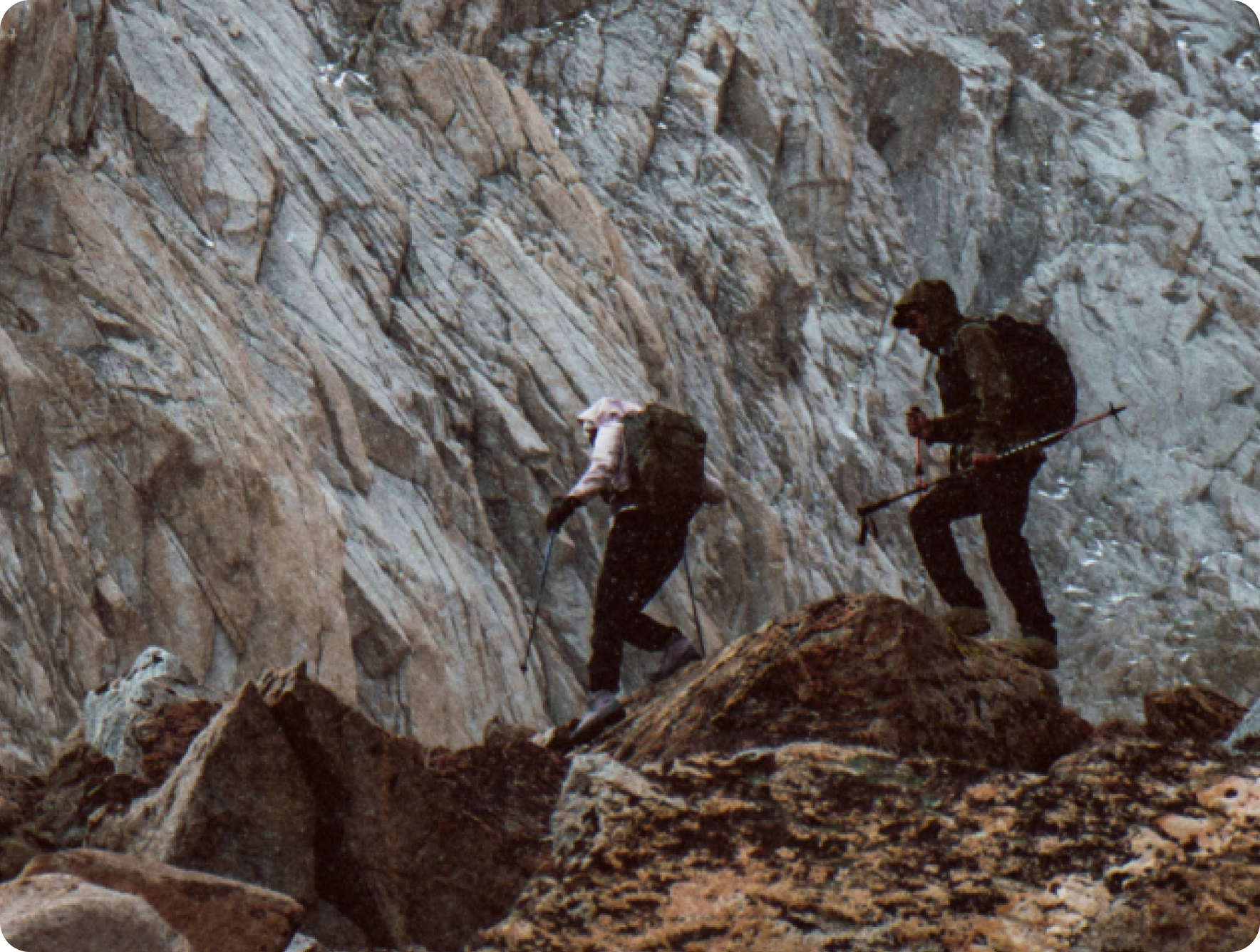 Two hikers with backpacks and trekking poles ascend a steep, rocky mountain slope.