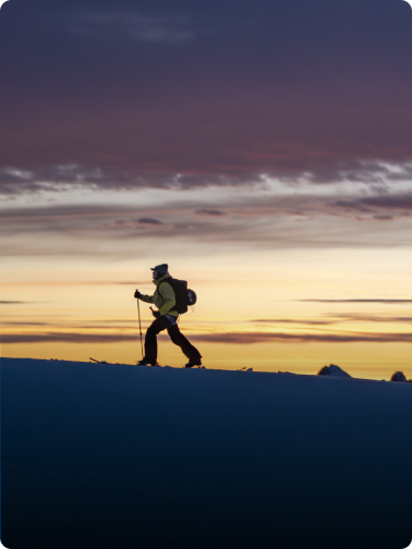 A person ski touring on a snowy ridge against a colorful sunset sky.