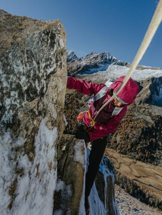 A climber in a pink jacket ascends a snowy rock face with a mountain range and autumnal trees in the background.