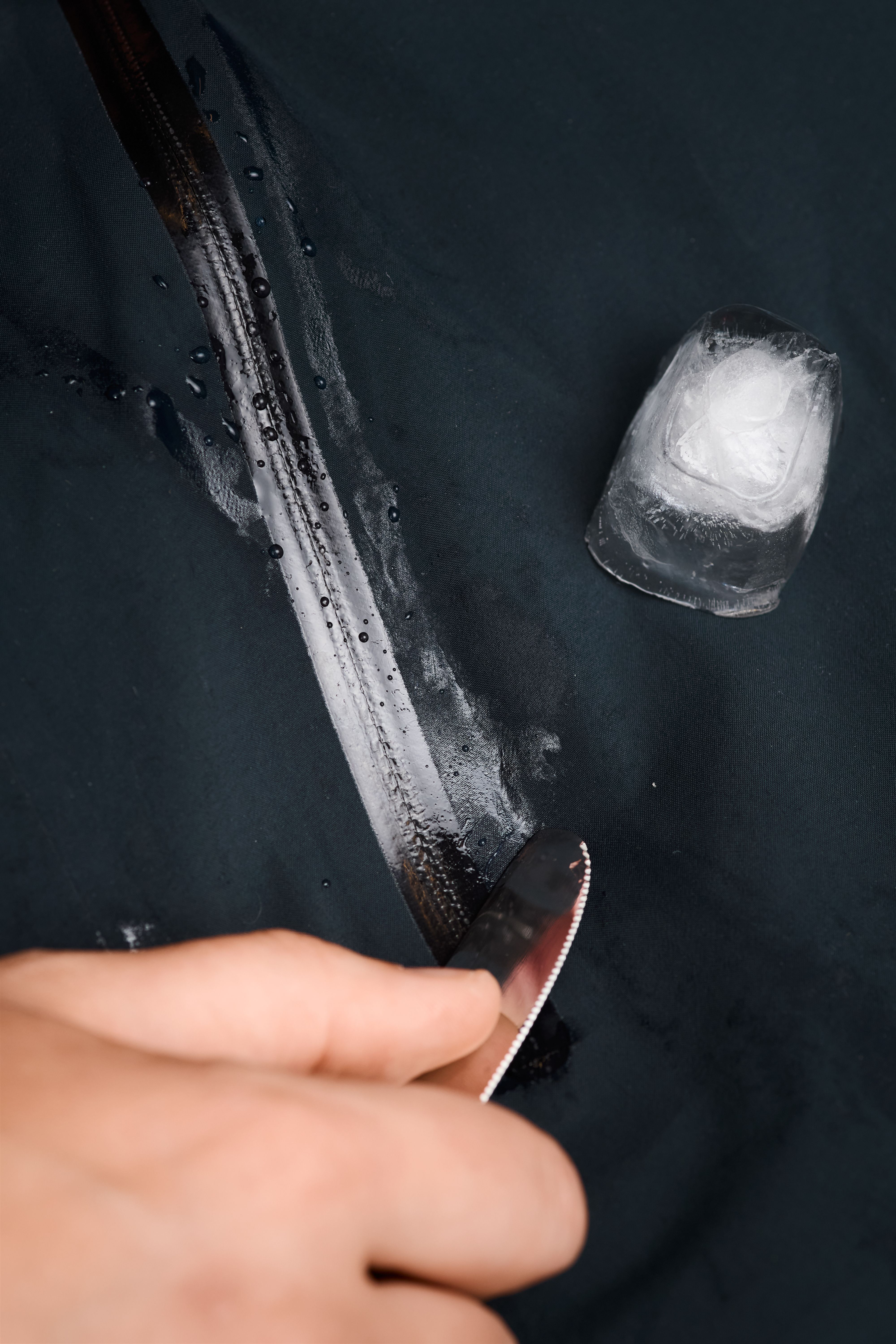 A hand scrapes a knife along dark waterproof fabric, leaving a trail of water next to a piece of ice.