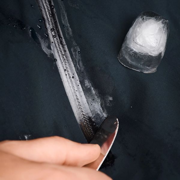 A hand scrapes a knife along dark waterproof fabric, leaving a trail of water next to a piece of ice.