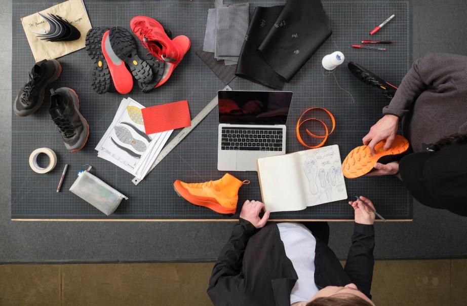 Two designers work on shoe prototypes at a table covered with components, sketches, and tools.