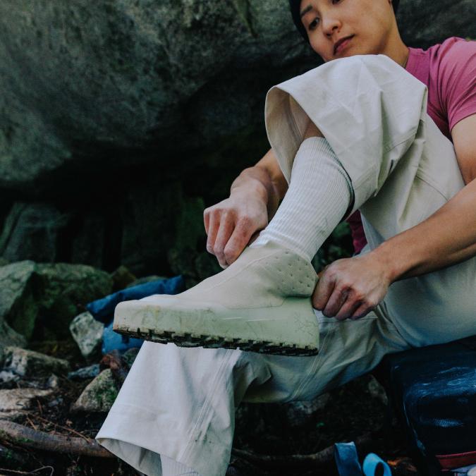 A person with chalk-dusted hands adjusts a beige slip-on outdoor shoe in a rocky environment.