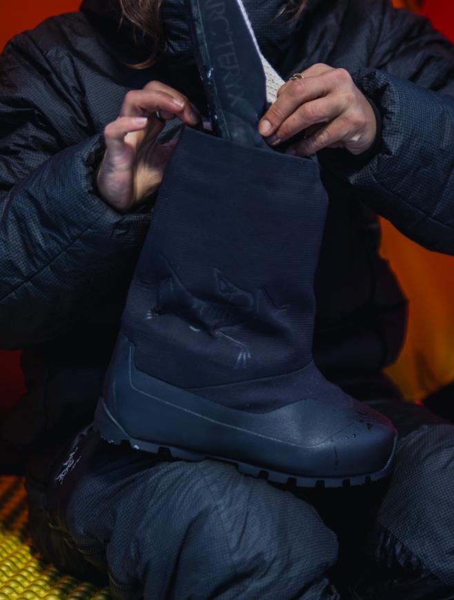 A person in a dark Arc'teryx jacket adjusts a black insulated boot.