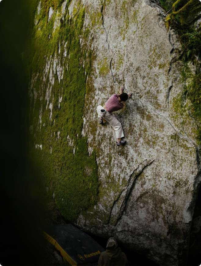 A person bouldering on a moss-covered rock with a spotter and crash pad below.