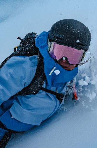 Overhead view of a skier in a black helmet, pink goggles, and blue jacket, kicking up snow.