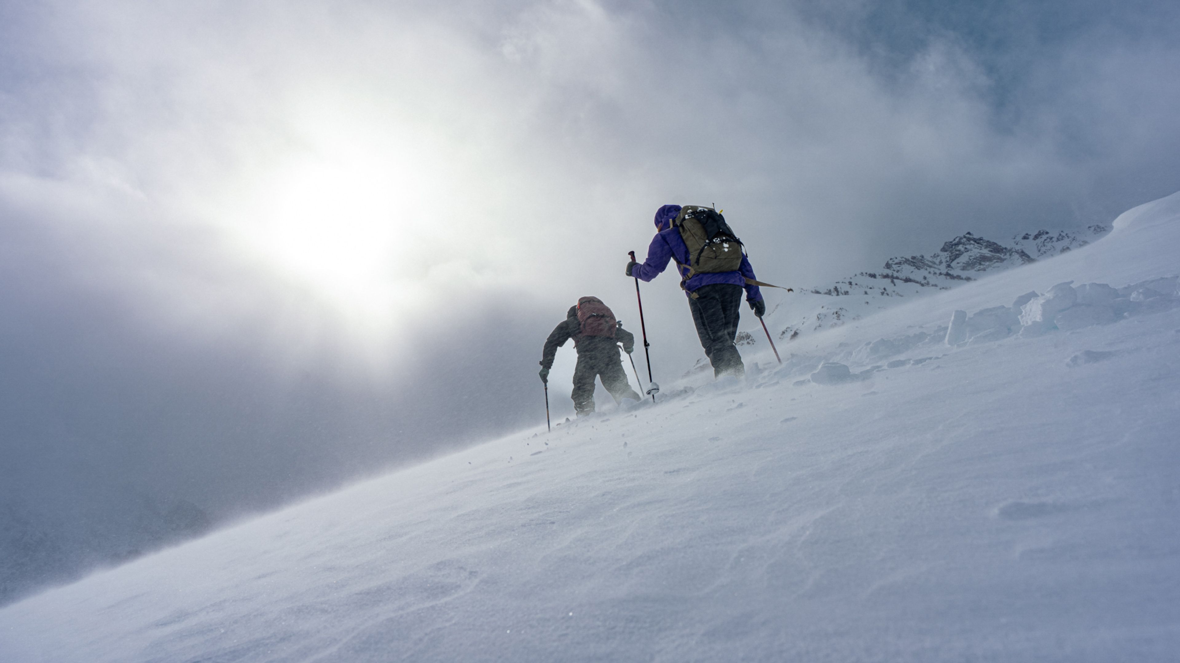 Two people ascend a snowy mountain slope with poles and backpacks under a bright, cloudy sky.