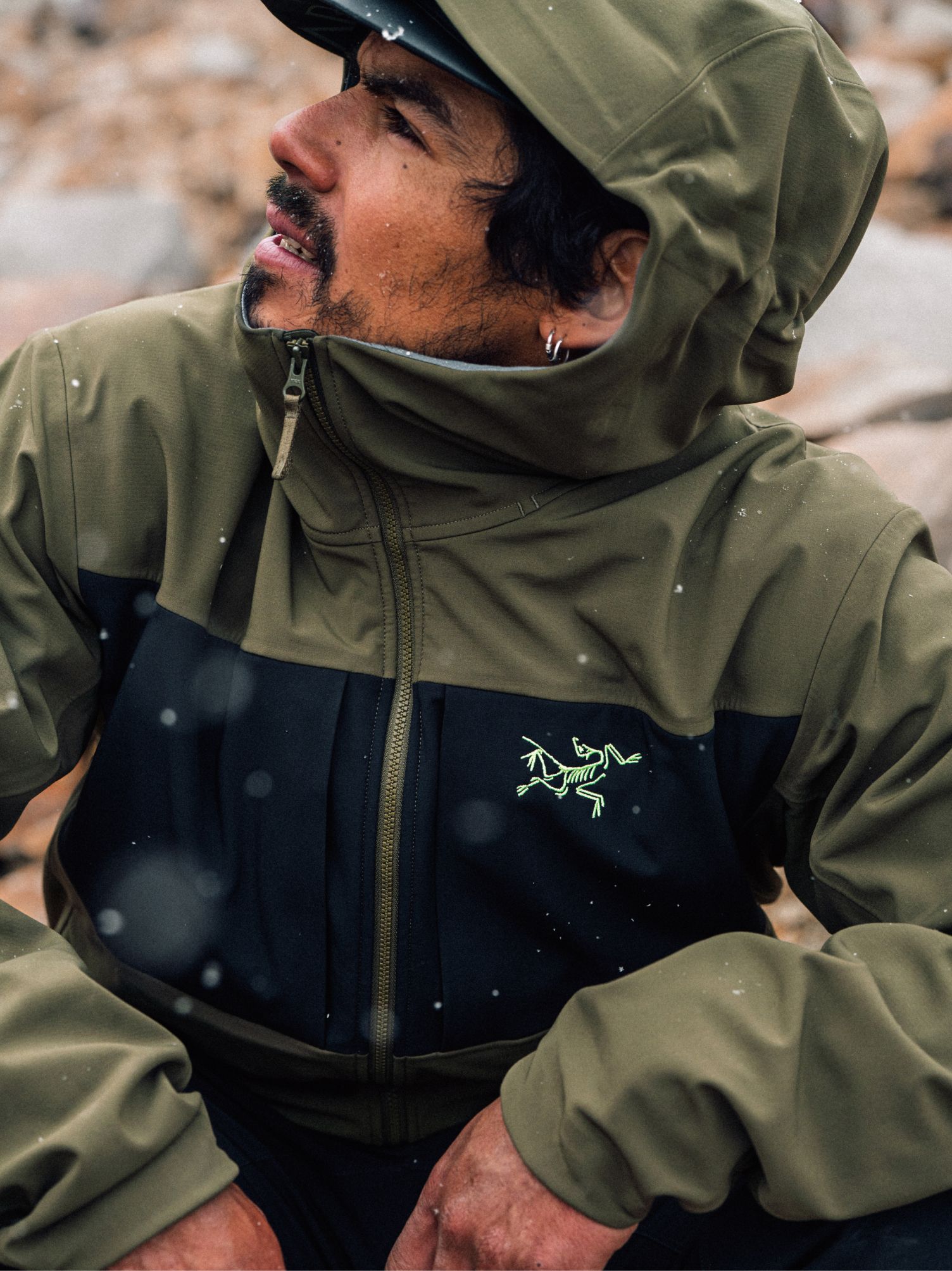 A man in an olive green and black Arc'teryx jacket, looking up as light snow falls.