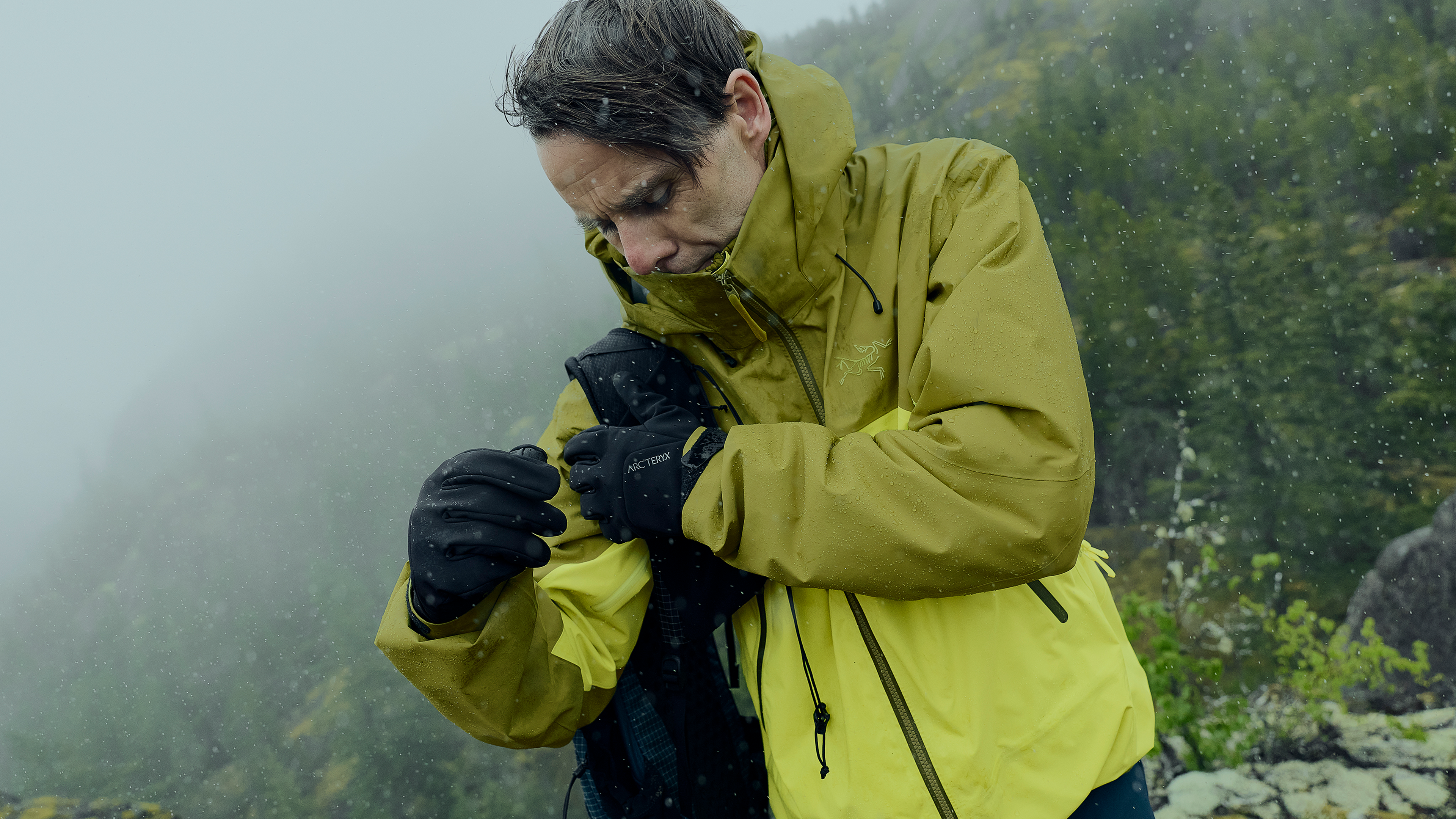 Person in a yellow-green rain jacket and gloves adjusting their gear in a snowy, foggy mountain environment.