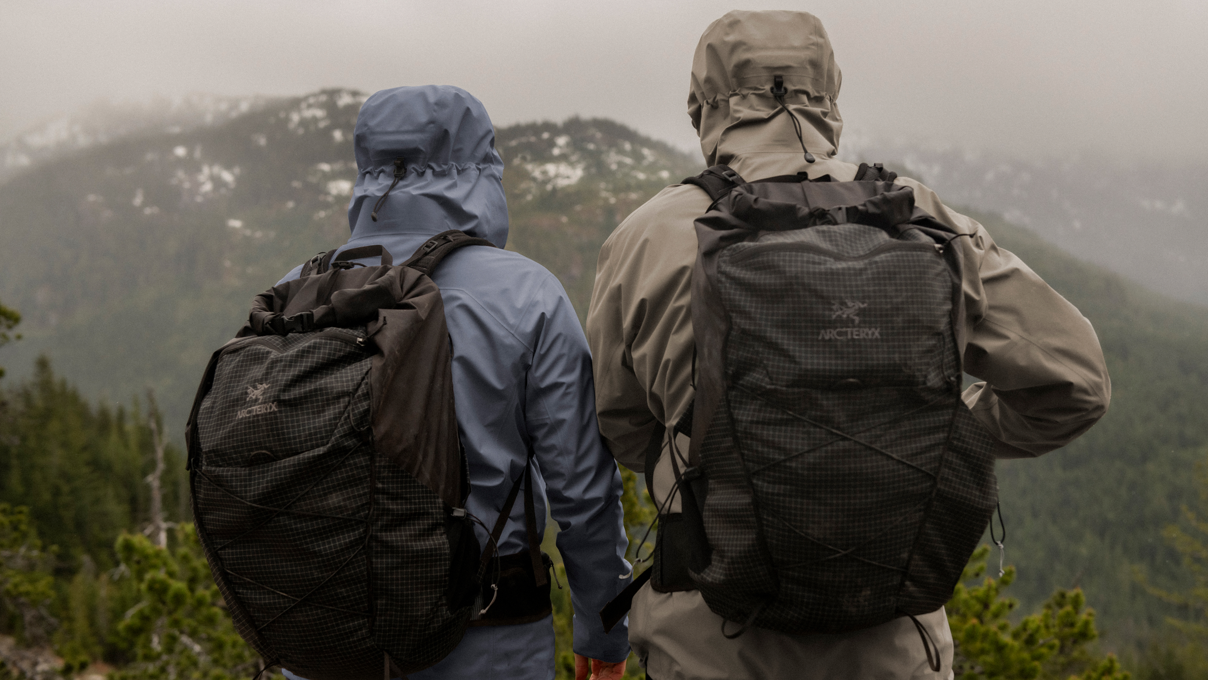 Two people in hooded jackets and Arc'teryx backpacks look out at a misty, forested mountain range.
