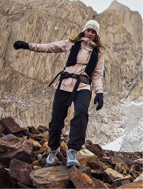 A female hiker in a pink jacket, dark pants, and white beanie carefully descends a rocky mountain path under light snow.