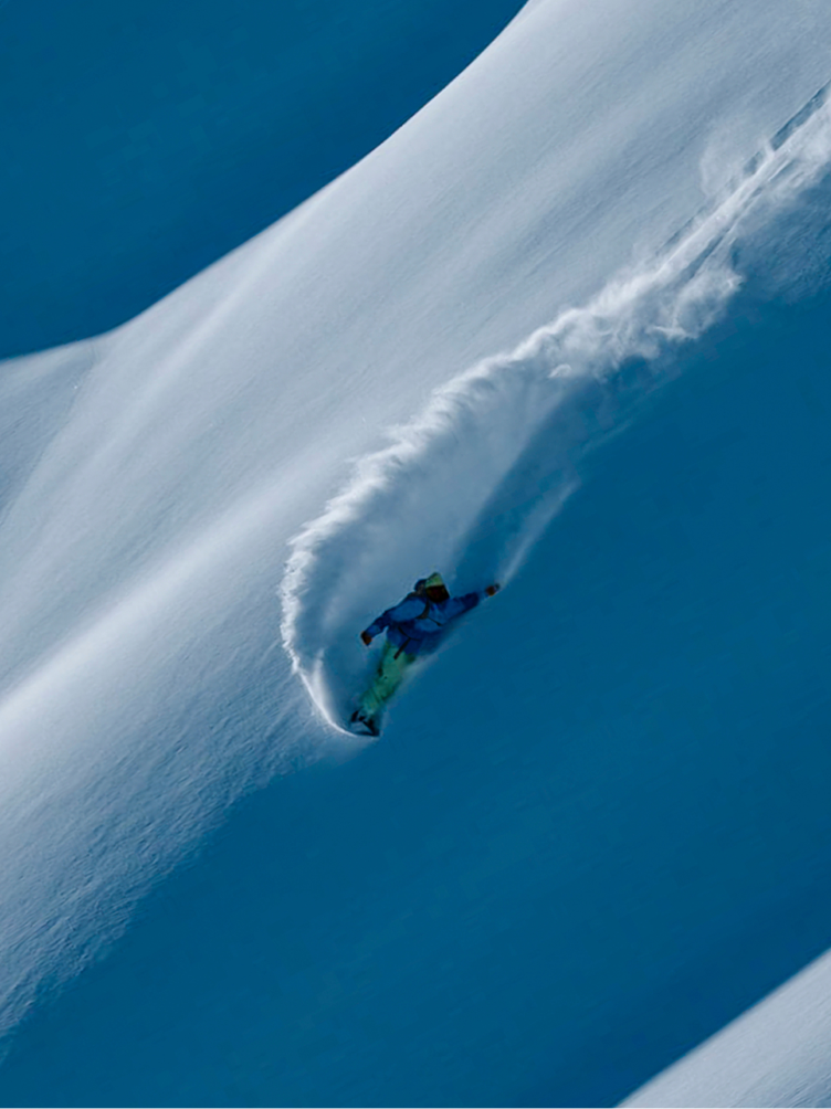 Snowboarder carving a turn on a steep, snowy slope, kicking up a trail of powder.