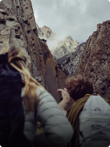 Two hikers gaze up at dramatic, cloud-covered rocky peaks.