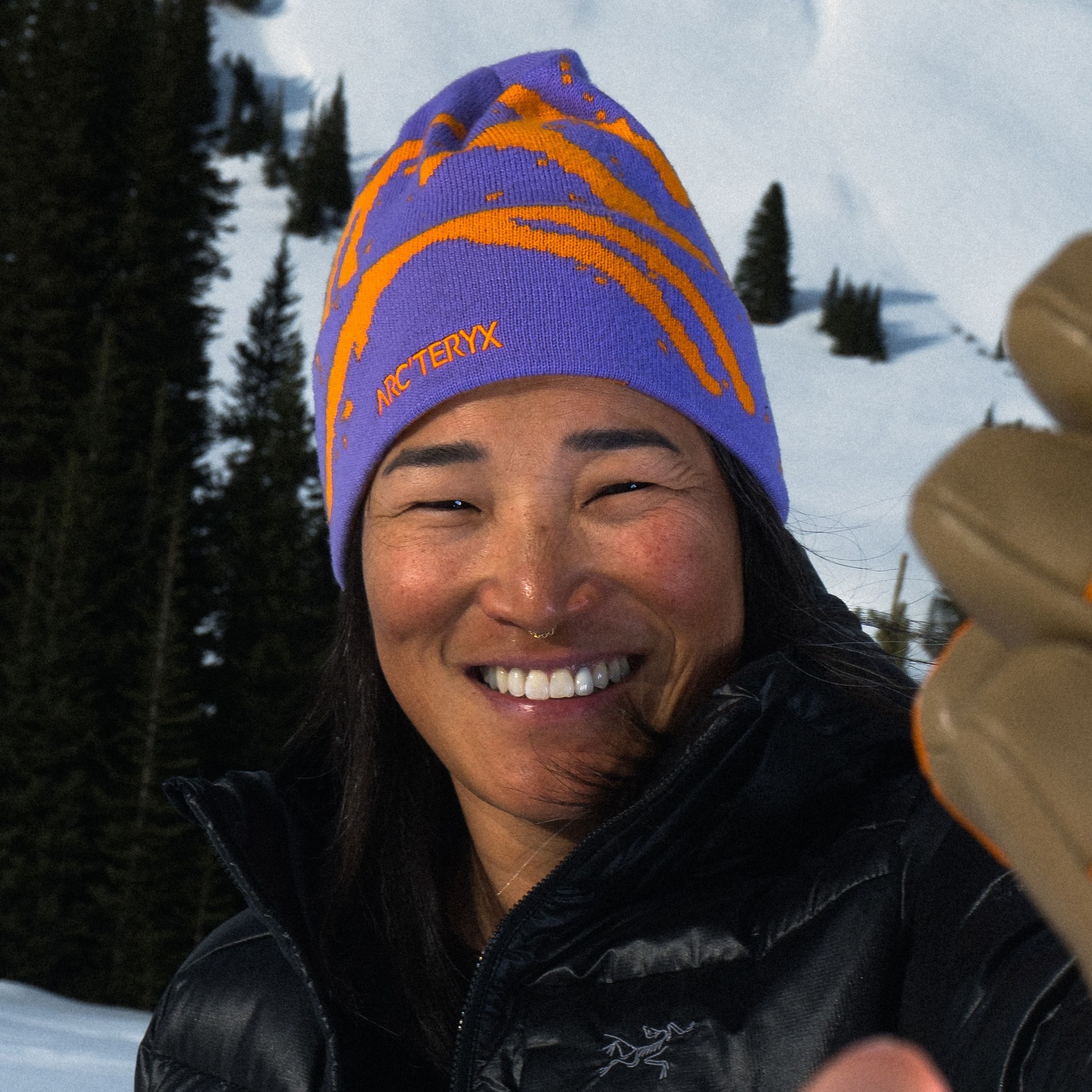 A smiling woman in a purple and orange Arc'teryx beanie, black jacket, and brown glove, in front of snowy mountains and trees.