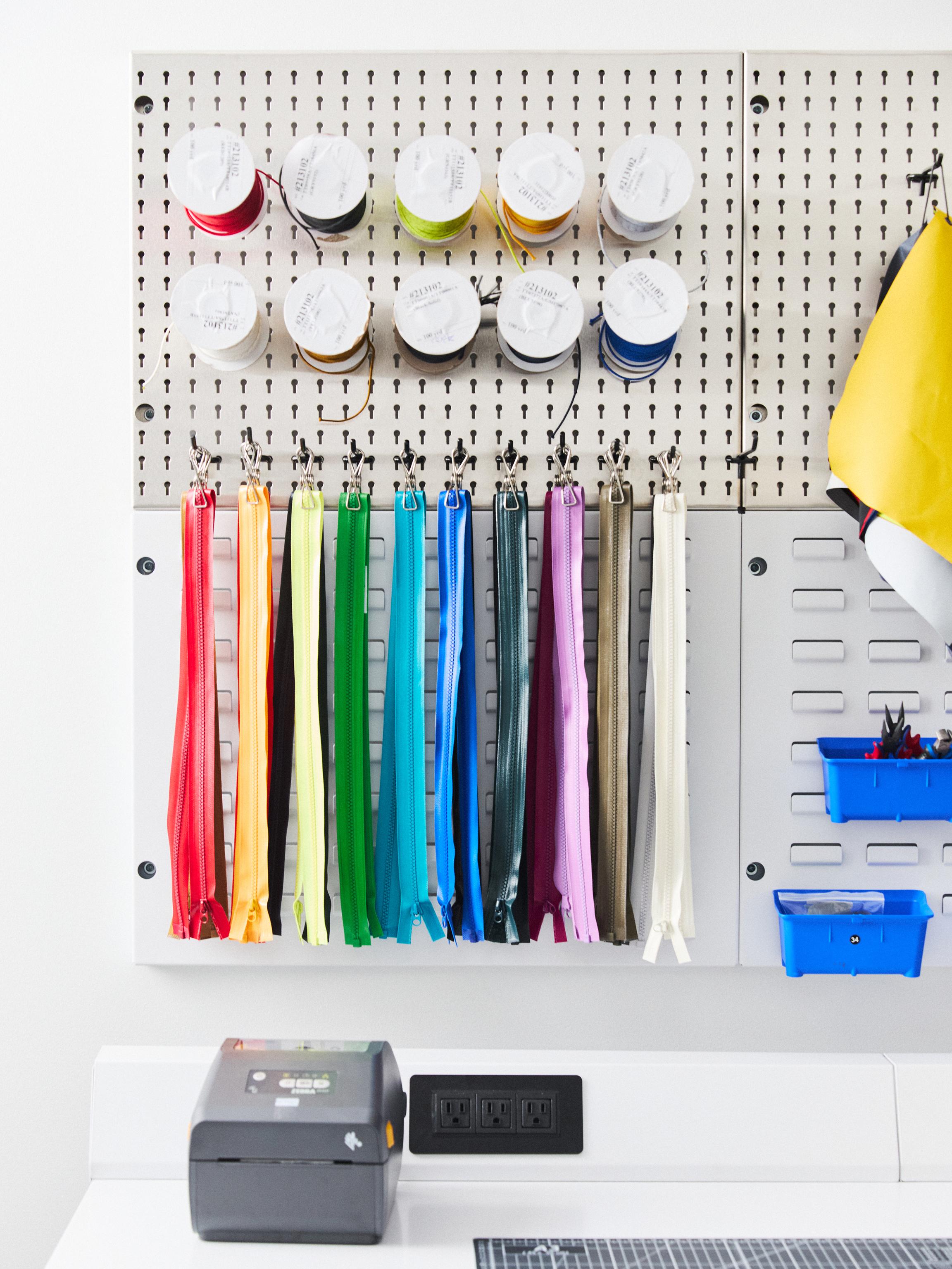 A craft organization system featuring a pegboard with spools of thread and colorful zippers, above a counter with a label printer, power outlet, and blue storage bins.