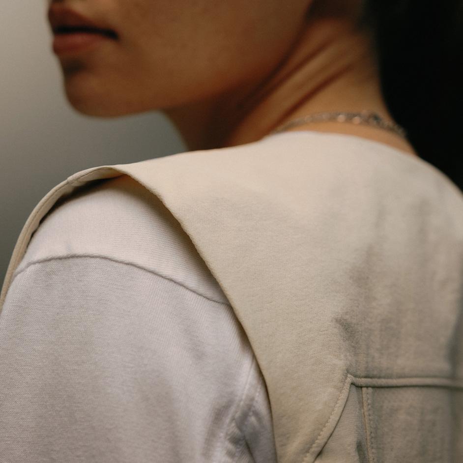Close-up of a person from behind, wearing a cream-colored garment with a wide shoulder flap over a white t-shirt, with a silver necklace visible.