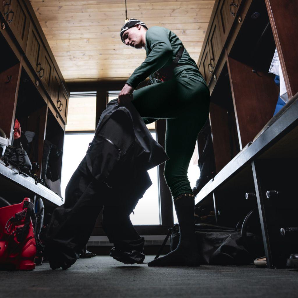 A person in green base layers puts on dark pants in a locker room.