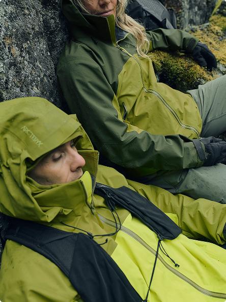 Two people in green and lime green Arc'teryx jackets resting against a mossy rock.