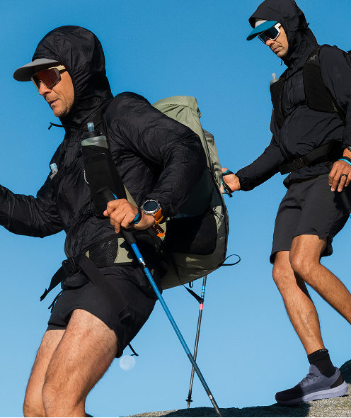 Two men in full hiking gear, including jackets, shorts, backpacks, and trekking poles, ascend rocky terrain.