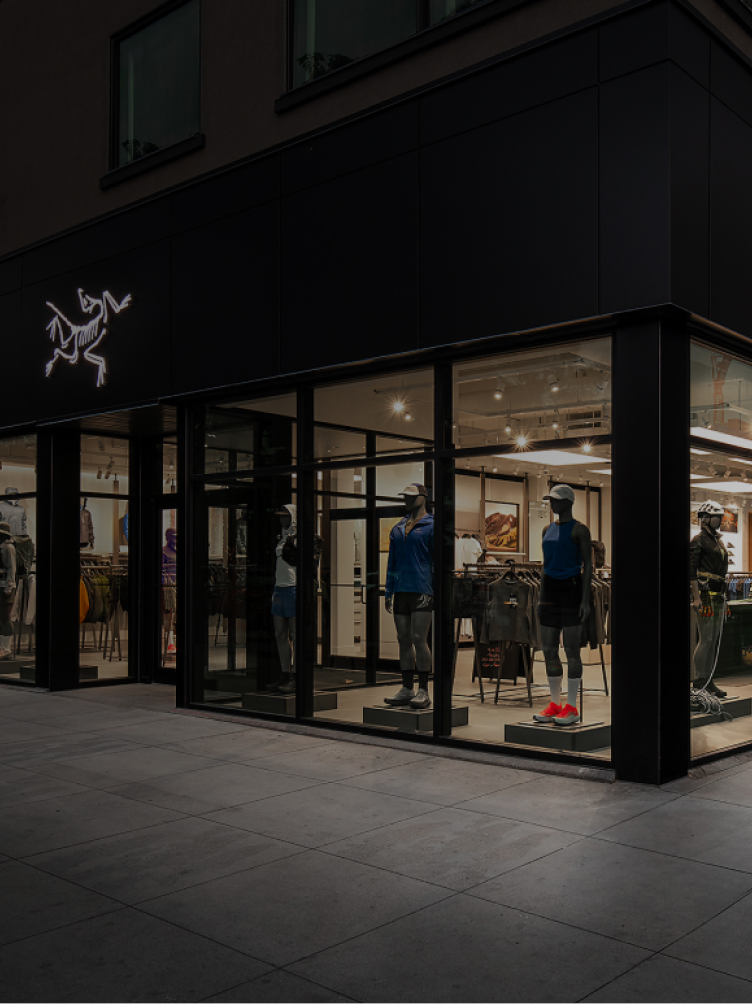 An Arc'teryx store at night with its illuminated logo and mannequins in activewear visible through large glass windows.