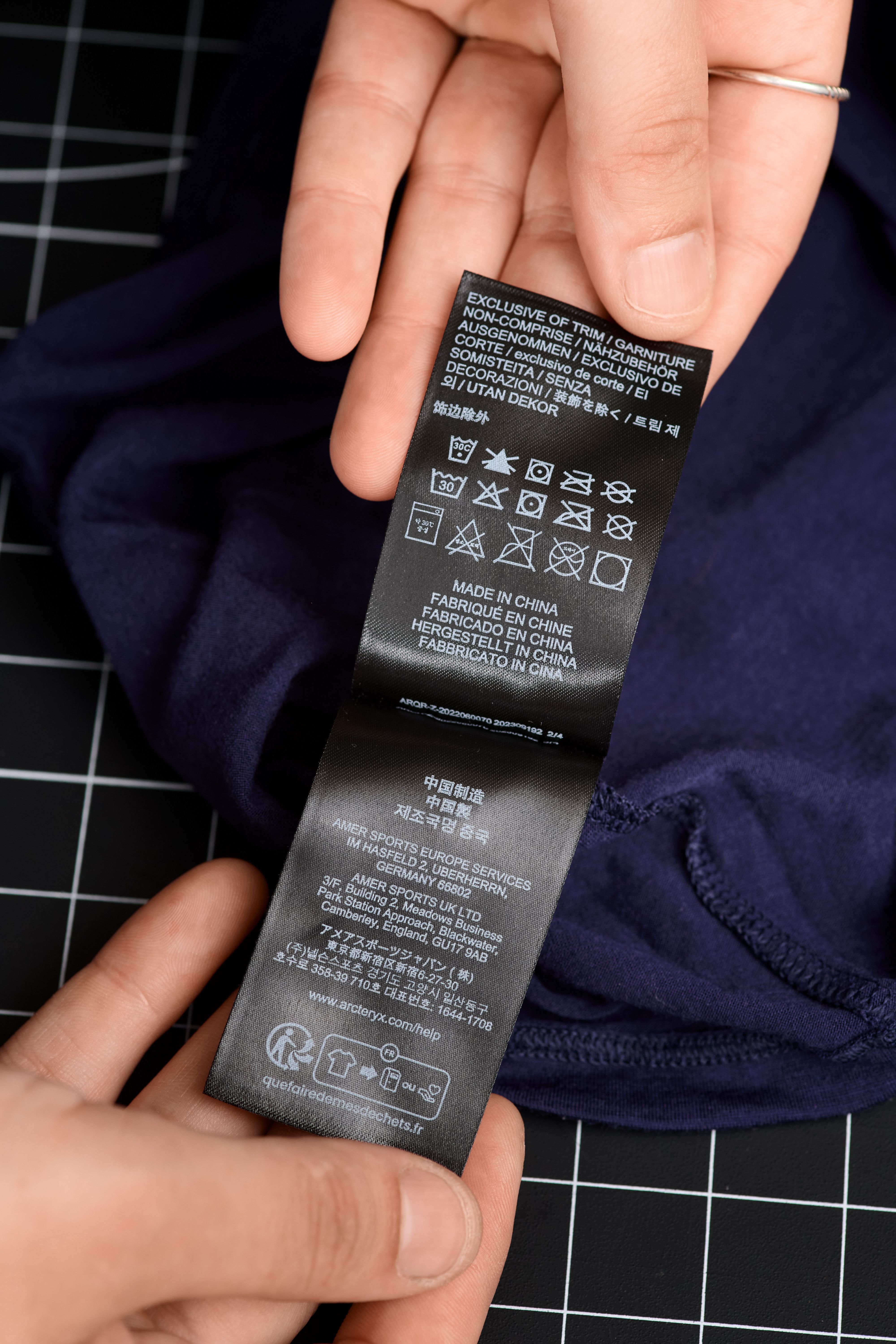 Hands hold a black clothing label with text and care symbols attached to a dark blue garment.