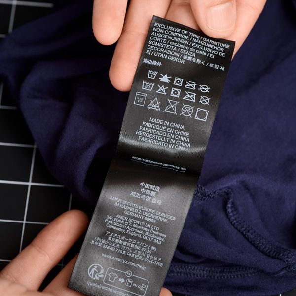 Hands hold a black clothing label with text and care symbols attached to a dark blue garment.
