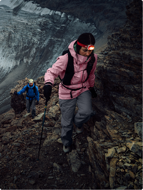 A woman in a pink jacket and headlamp ascends a rocky mountain trail, followed by a man, with a glacier behind them.