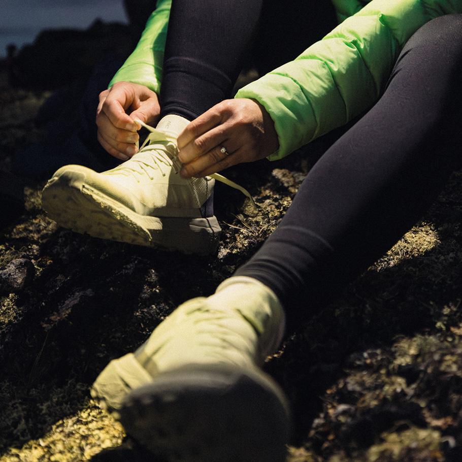 Person in a green jacket and black leggings tying a light green trail shoe on dark ground.