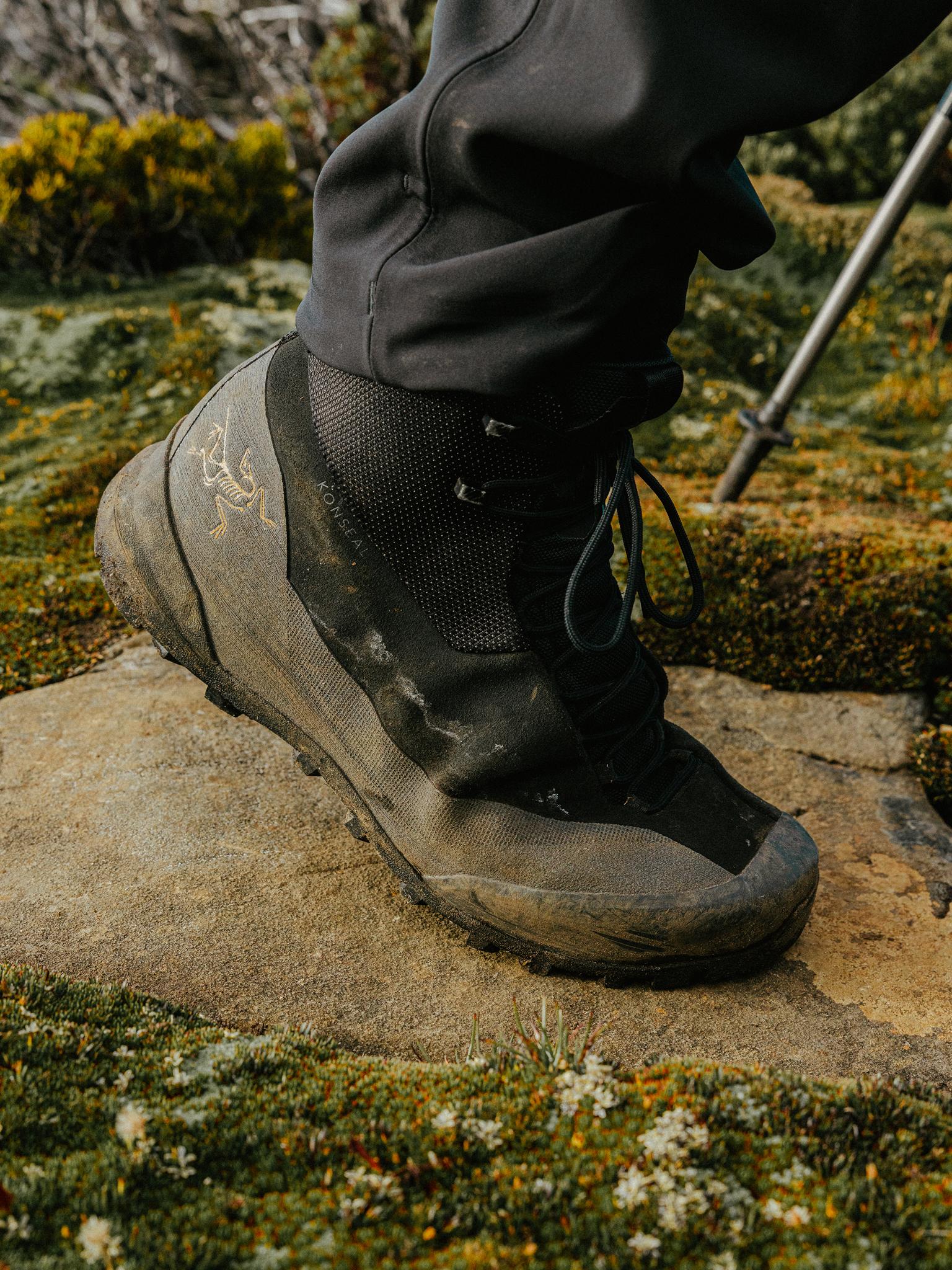 Close-up of a dirty Arc'teryx Konseal hiking boot on a rock amidst mossy terrain.