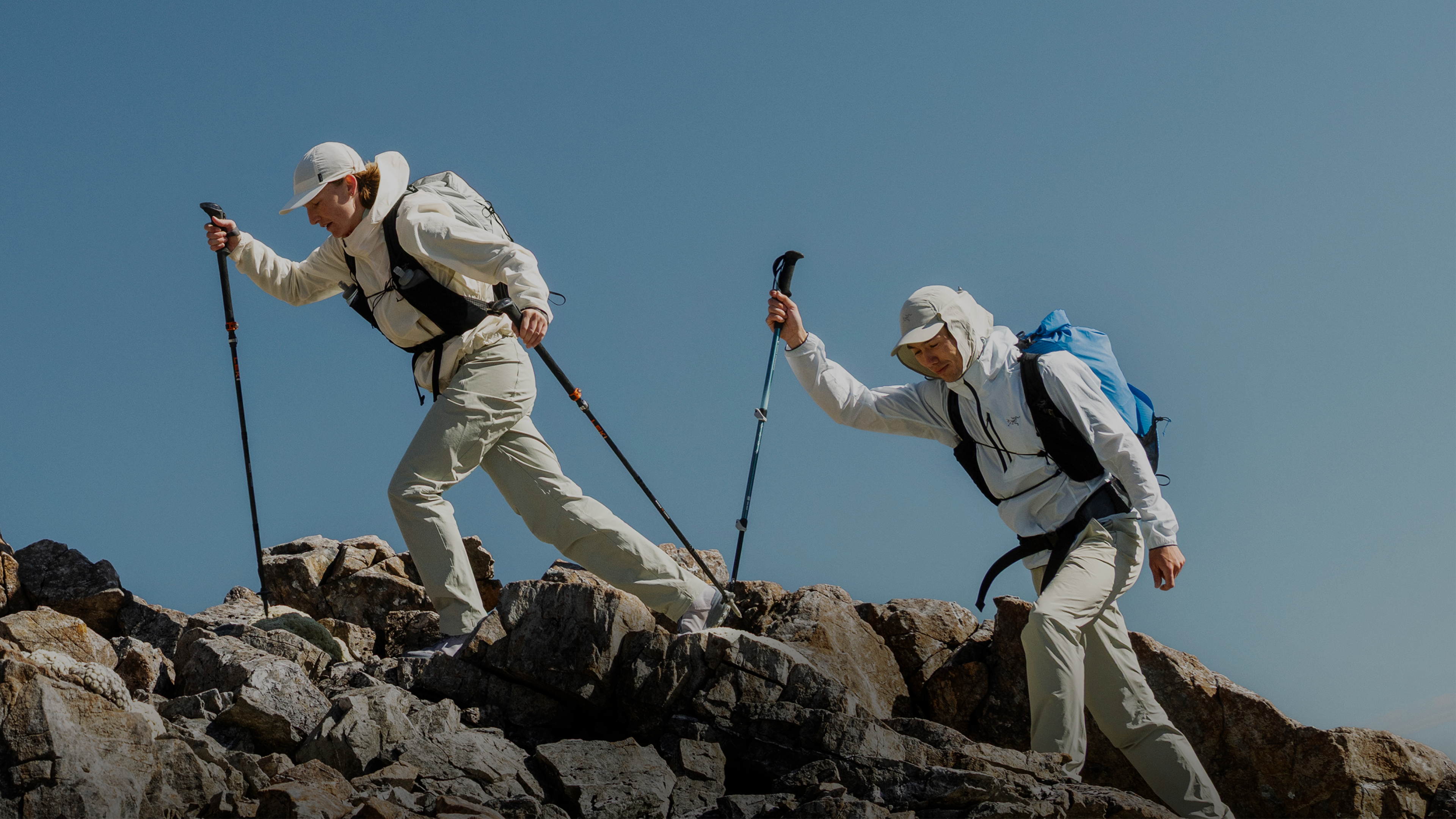 Two hikers ascend a rocky mountain path with trekking poles under a clear blue sky.