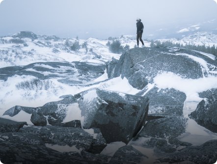 A person stands on a large, snow-dusted rock in a vast, wintry landscape.